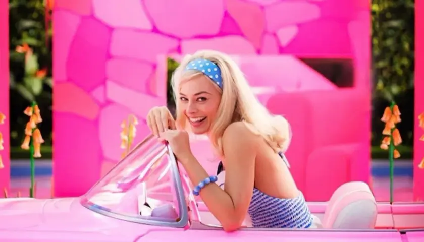 Margot Robbie jako Barbie w różowym kabriolecie, z uśmiechem pozuje na tle jaskrawego, różowego świata.