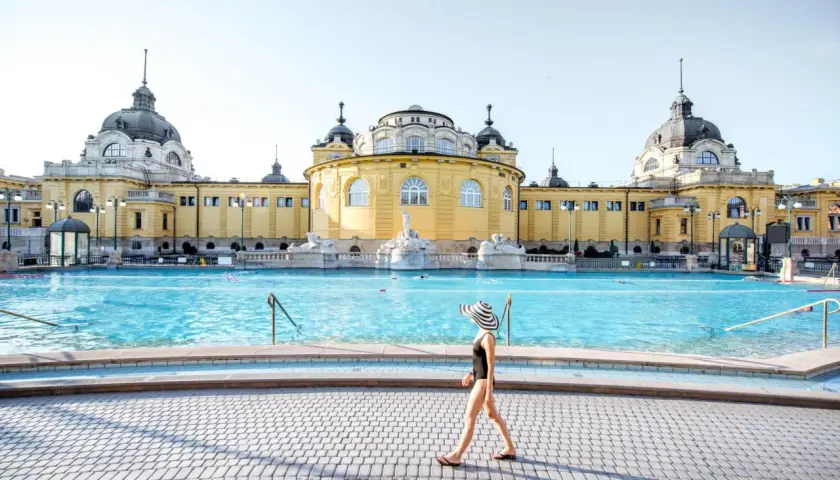 Frau mit Hut spaziert am Beckenrand des Budapester Thermalbads. Das beeindruckende gelbe Gebäude mit Kuppeln bildet die Kulisse.