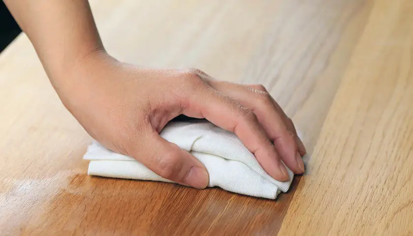 Dłoń z białym materiałem wciera olej w drewniany blat, odnawiając jego powierzchnię.