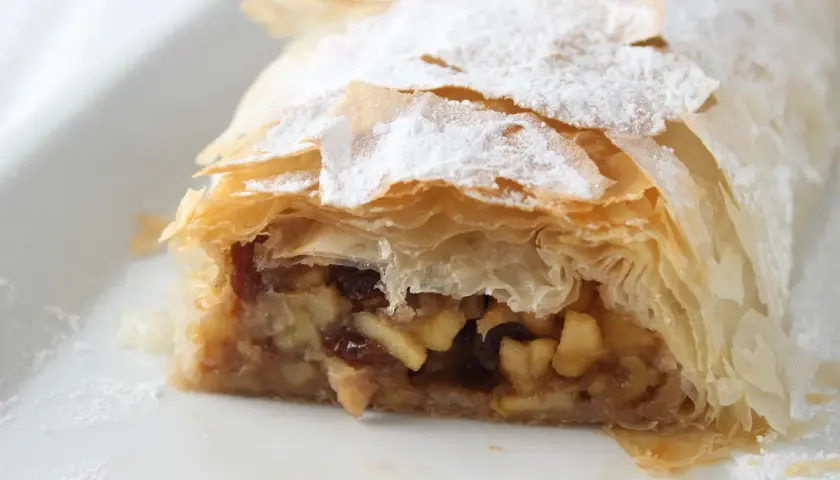 Un trozo de strudel de manzana, con su masa hojaldrada y relleno de manzana y pasas, espolvoreado con azúcar glas.