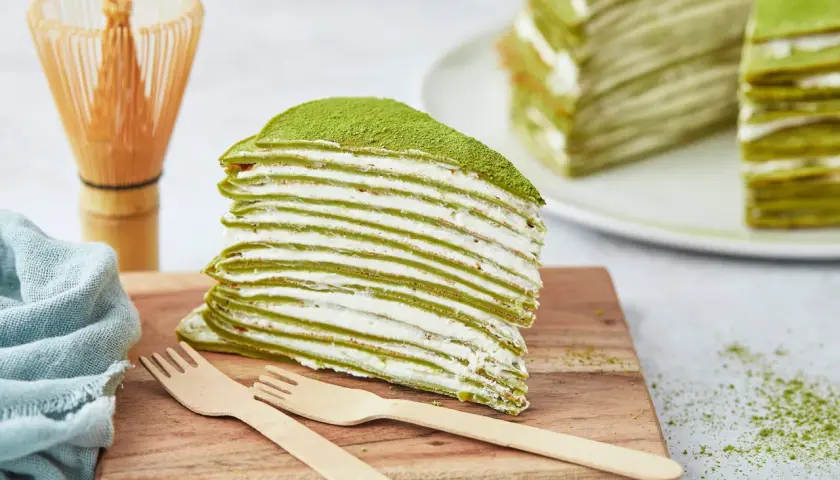 Ein Stück saftiger Matcha Kuchen mit vielen dünnen Schichten und cremiger Füllung, bestäubt mit grünem Pulver. Daneben ein Chasen und Holzgabeln.