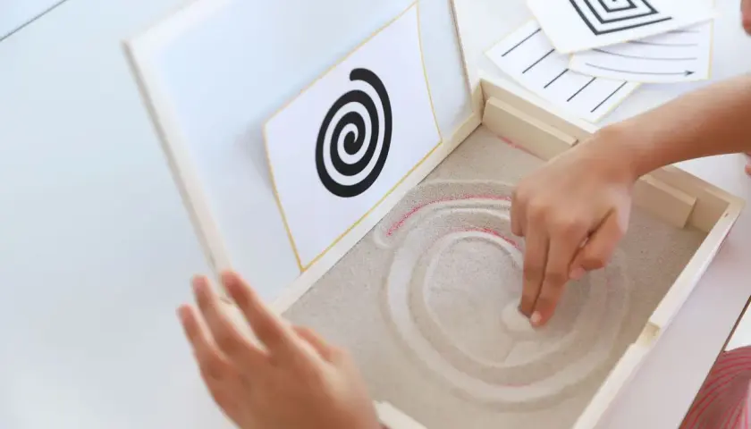 Niño practicando preescritura montessori, trazando espirales en arena.