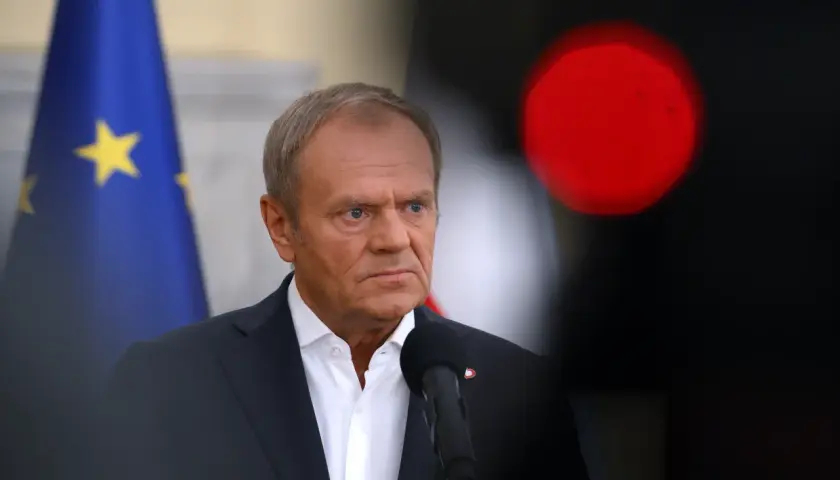 Donald Tusk w garniturze, z flagą UE w tle i czerwonym światłem.