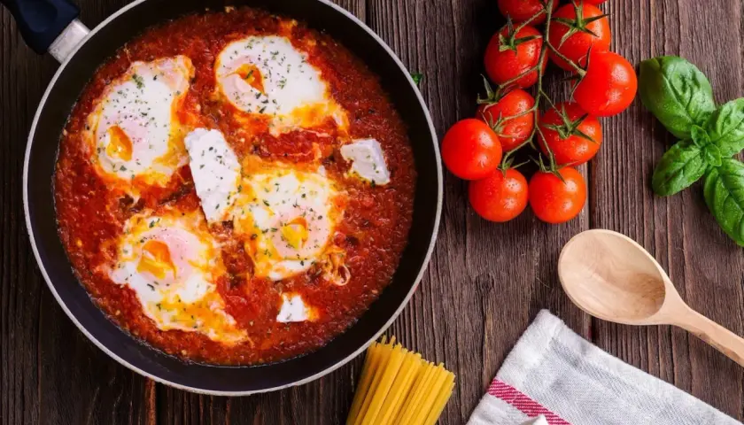 Uova al pomodoro in padella, con pomodorini freschi, basilico e un cucchiaio di legno, pronte per essere gustate.
