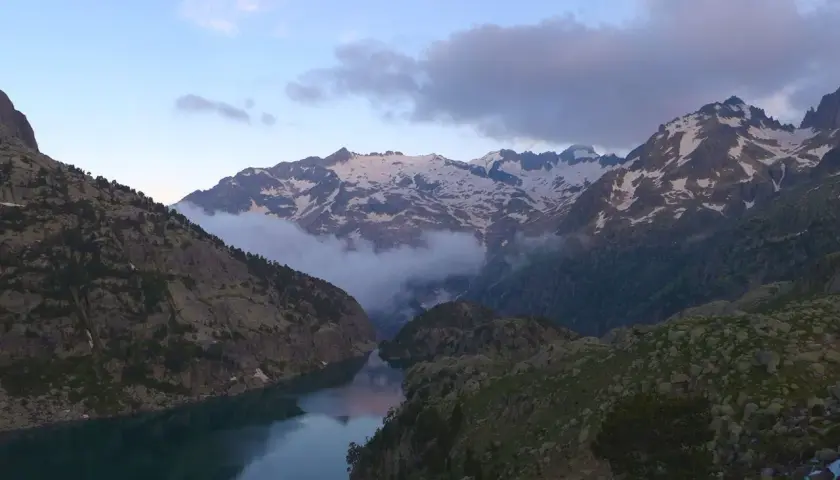 Paisaje montañoso con un lago sereno y picos nevados. Nubes bajas envuelven las montañas, creando una atmósfera mística. Un lugar idílico para explorar, como el que se encuentra en Besiberri Sud.