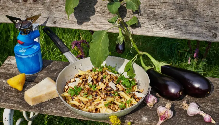 Pasta melanzane mit frischen Auberginen, Knoblauch und Parmesan. Ein Campingkocher und Zitrone deuten auf ein Outdoor-Mahl hin.