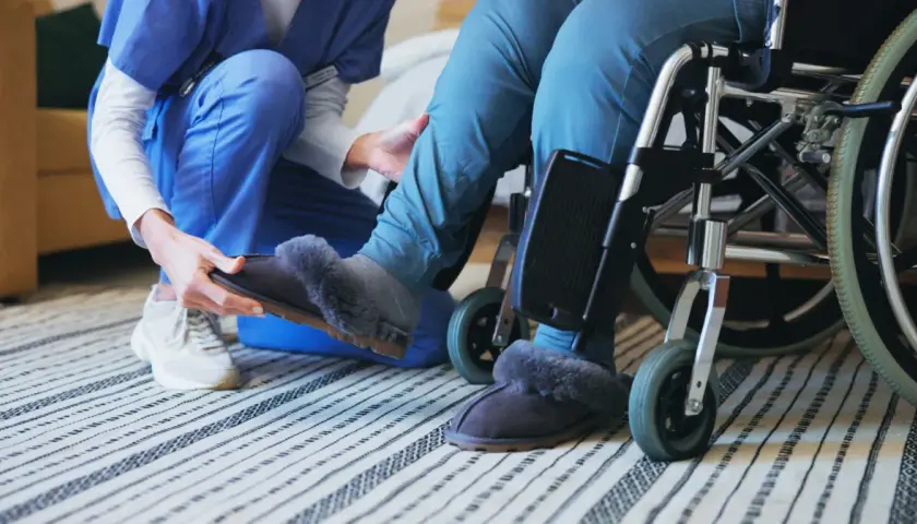 Opiekunka zakłada kapcie osobie na wózku. To codzienne świadczenie wspierające, pełne troski i empatii.