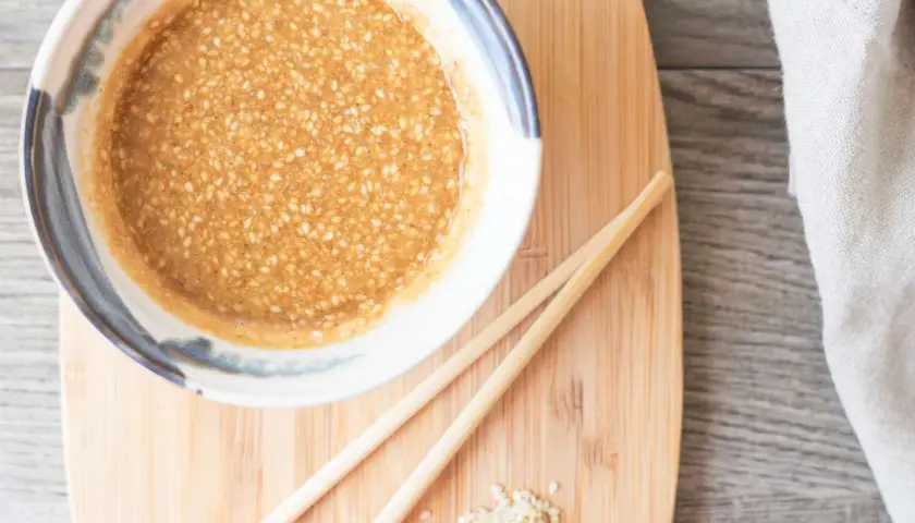 A bowl of creamy sesame sauce, perfect for your next sesame sauce recipe, sits on a wooden board with chopsticks and a sprinkle of sesame seeds.