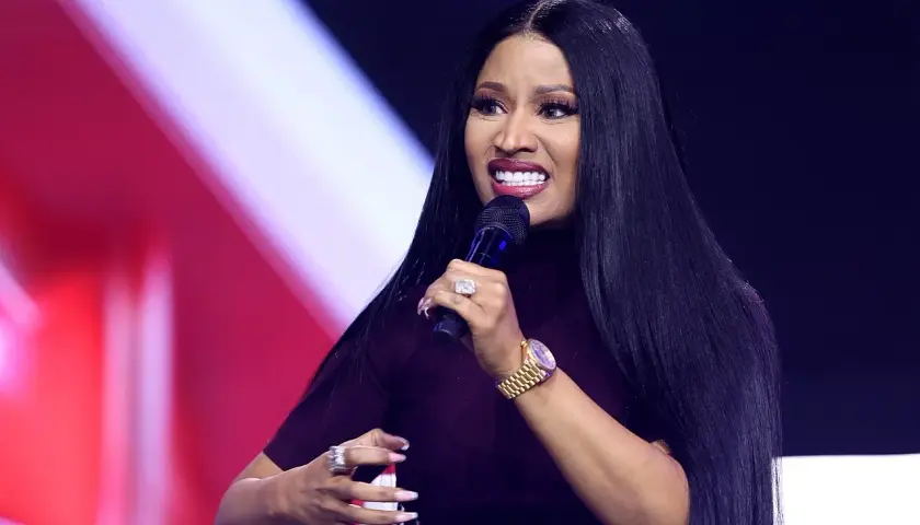 Nicki Minaj z mikrofonem, w ciemnej sukience, z długimi czarnymi włosami i biżuterią.