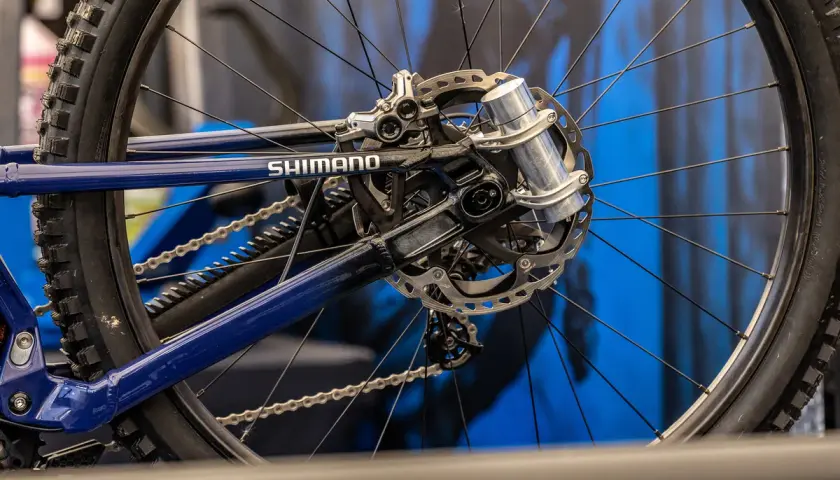 Close-up of a blue bicycle's rear wheel and Shimano drivetrain. A silver grip damper is visible on the axle, ready to absorb shocks.