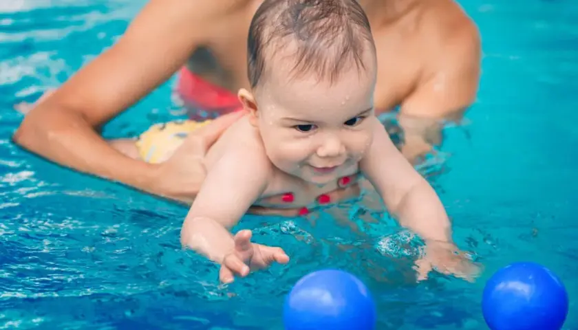 Glad bebis med ett leende i vattnet, en person håller i den. Två blå bollar flyter framför. Perfekt för babysim ålder.