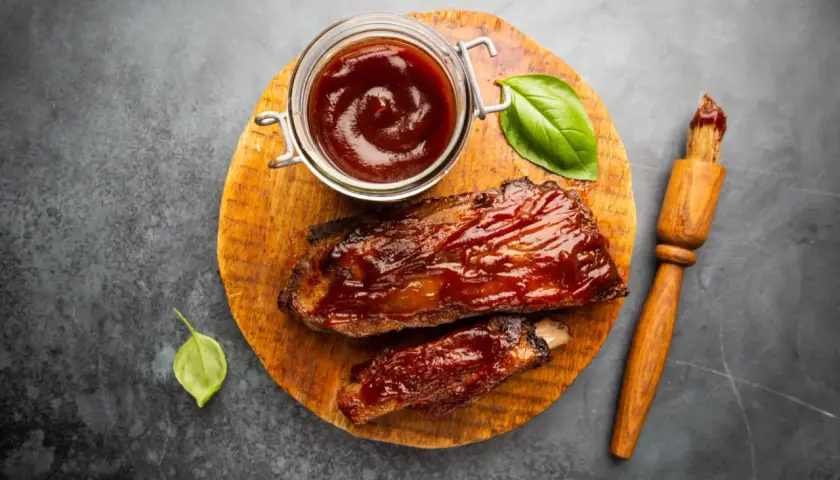 Soczyste żeberka w sosie barbecue, gotowe do podania. Obok słoiczek z sosem do steka i pędzelek.