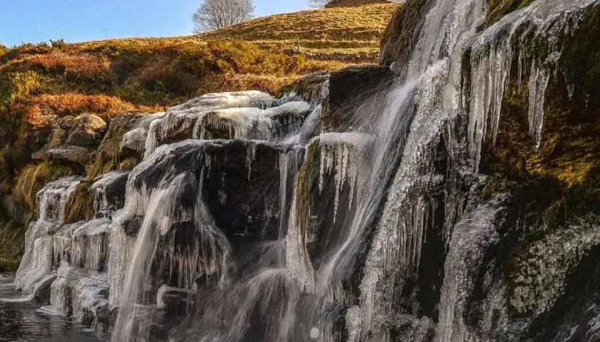 Cascada helada en los valles pasiegos, con el sol brillando sobre la ladera y el cielo azul.