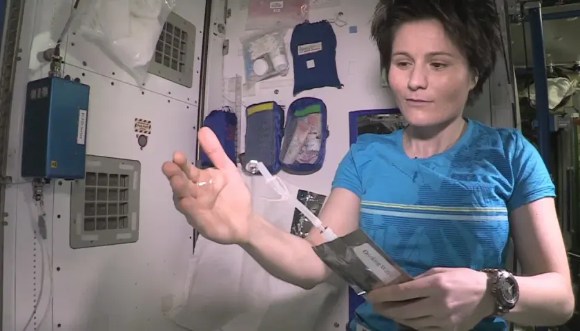 Astronautka pokazuje kroplę wody w dłoni, która unosi się w przestrzeni kosmicznej. Trzyma też opakowanie z napisem "Drinking Water".