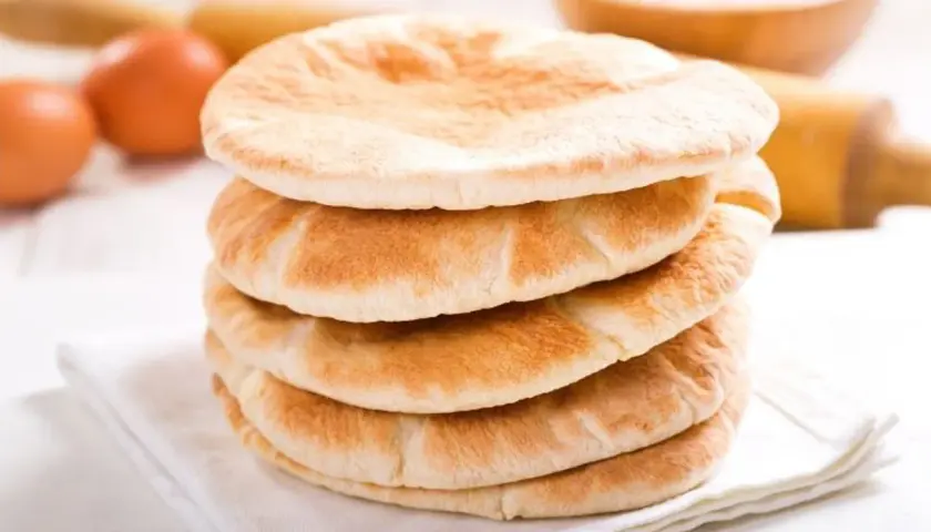 Stos świeżo upieczonego chleba arabskiego na białej serwetce, obok jajka i wałka do ciasta.