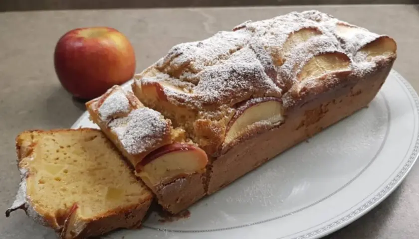 Plumcake alle mele soffice e profumato, cosparso di zucchero a velo, con fette di mela a vista. Una mela rossa intera sullo sfondo.