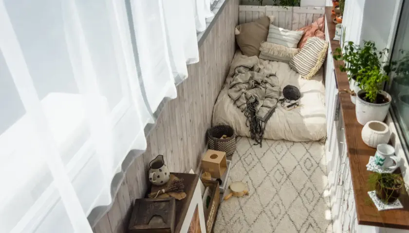 Przytulny balkon w bloku z materacem, kocem i poduszkami. Dywanik i rośliny dodają uroku.