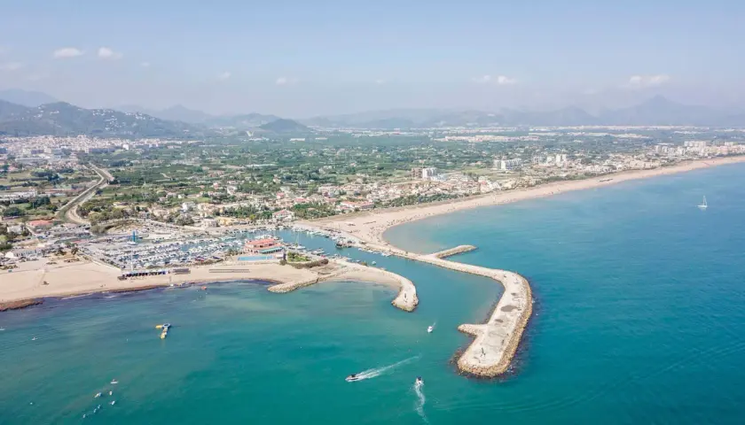 Vista aérea del puerto de Oliva con barcos navegando y la playa llena de gente.