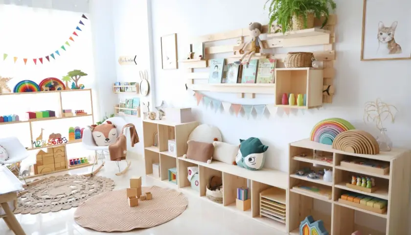 Un rincón Montessori lleno de juguetes de madera, arcoíris y peluches. Estanterías de pino organizan libros y materiales educativos.