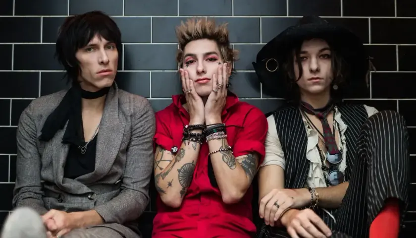 Trzech członków Palaye Royale w stylowych, rockowych strojach siedzi obok siebie na tle czarnych płytek.