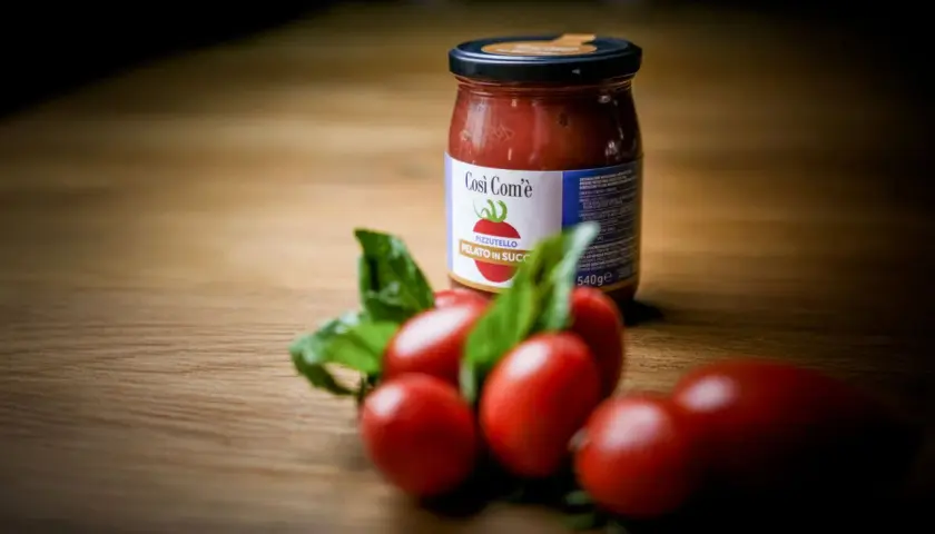 Barattolo di pomodoro siccagno pizzutello pelato in succo, con pomodorini freschi e basilico su un tavolo di legno.