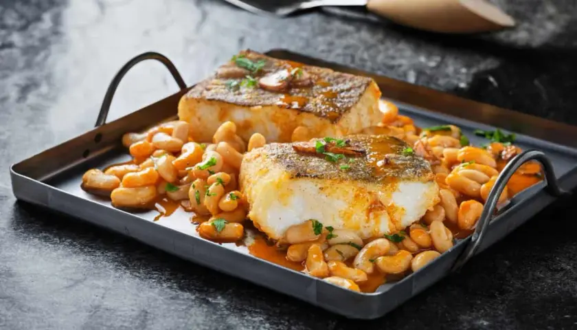 Dos lomos de bacalao a la llauna, dorados y jugosos, reposan sobre una cama de alubias blancas en salsa.