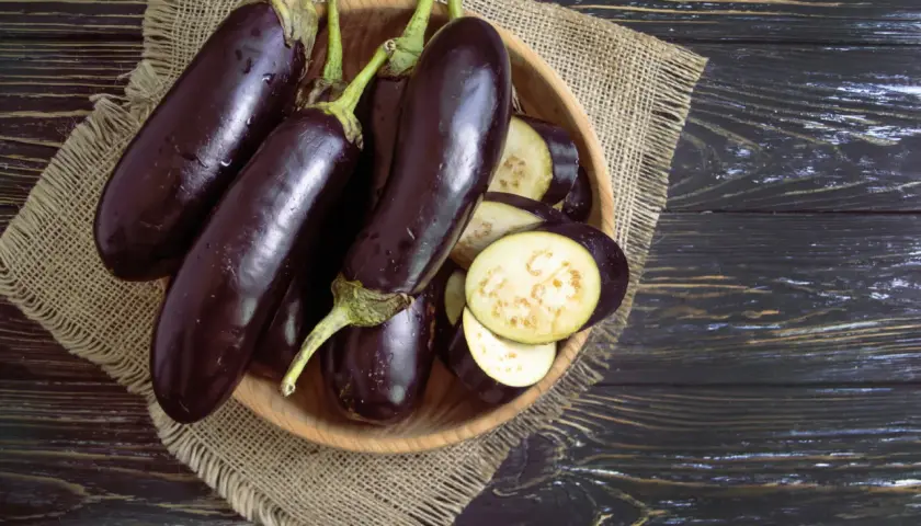 Einige ganze Auberginen liegen neben geschnittenen Scheiben in einer Holzschale. Man sieht das helle Fruchtfleisch der Aubergine innen.