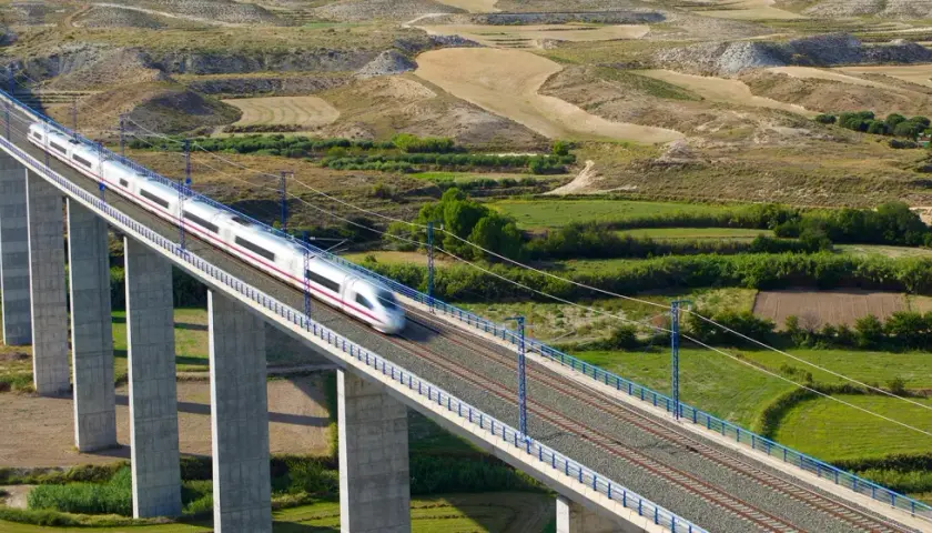 Un tren de alta velocidad cruza un viaducto sobre un paisaje rural. El tren, que parece ir de Lleida a Madrid, es plateado con una franja morada.