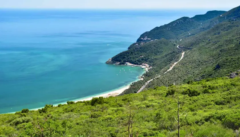 Costa verde en el sur de Lisboa, con mar turquesa, playa, barcos y montañas cubiertas de vegetación.