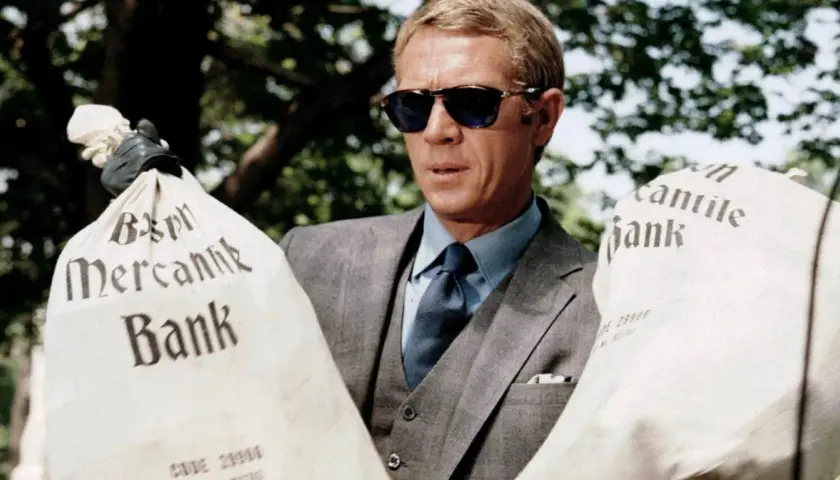 Steve McQueen w okularach przeciwsłonecznych, w eleganckim garniturze, trzyma worki z napisem "Mercantile Bank". Scena z filmu "Afera Thomasa Crowna".