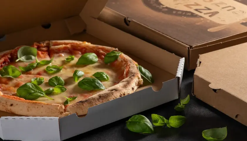 Pyszna pizza w otwartym opakowaniu na pizze, ozdobiona świeżą bazylią. Obok zamknięte kartony.