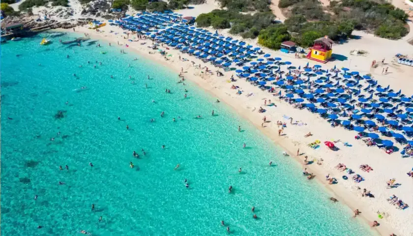 Słoneczny dzień na plaży Makronissos. Turkusowa woda, biały piasek, rzędy niebieskich parasoli i mnóstwo plażowiczów cieszących się słońcem i kąpielą.