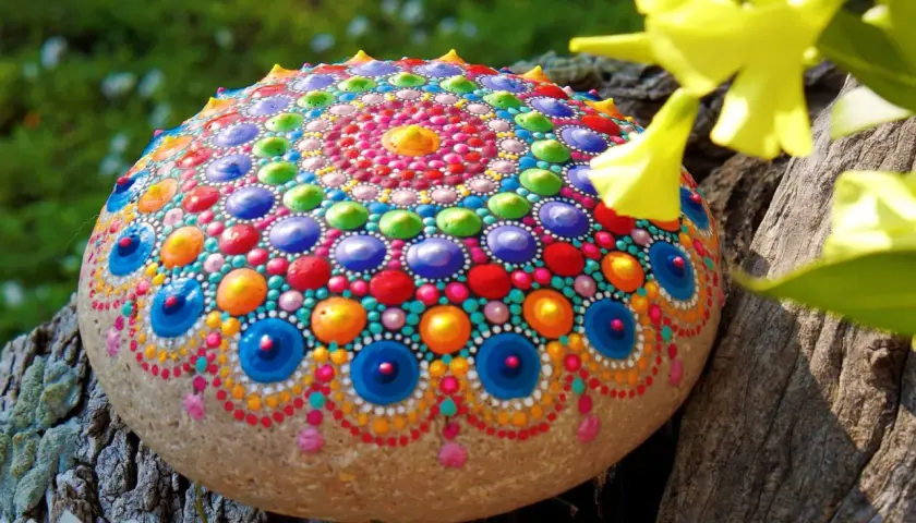 Bunte bemalte Steine mit Mandala-Muster, die auf einem Baumstamm liegen, umgeben von grünem Gras und gelben Blüten.