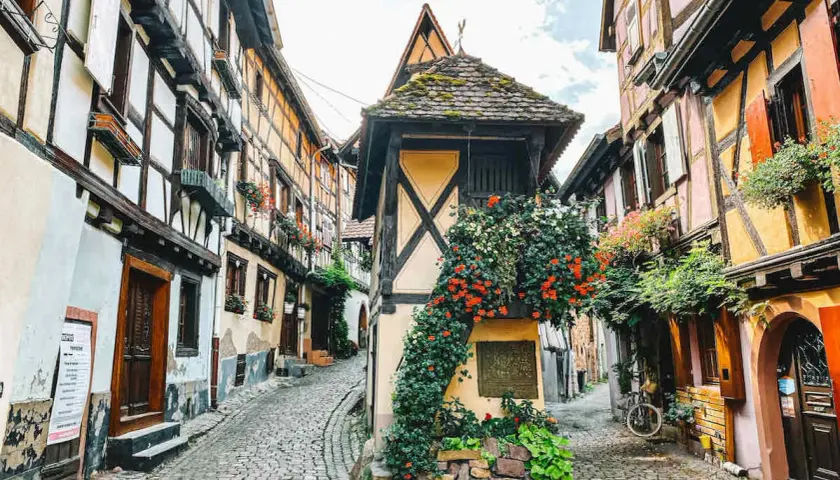 Kopfsteinpflastergasse im Elsass mit Fachwerkhäusern, Blumen und einem Fahrrad. Ein perfekter Ort für Geheimtipps im Elsass.
