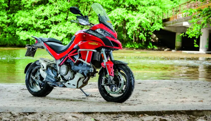 Czerwony Ducati Multistrada 1200S zaparkowany na piaszczystym brzegu rzeki, otoczony bujną zielenią.