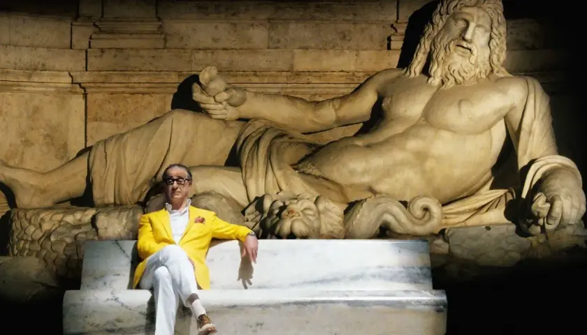 Un uomo in giacca gialla seduto su una panchina di marmo, con una statua antica alle spalle. Un'immagine che evoca la grande bellezza, spiegazione visiva dell'arte e del tempo.