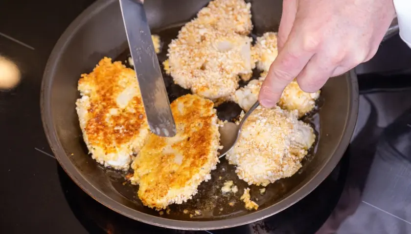 Goldbraune Blumenkohl-Schnitzel brutzeln in der Pfanne. Eine Hand wendet sie mit Messer und Löffel.