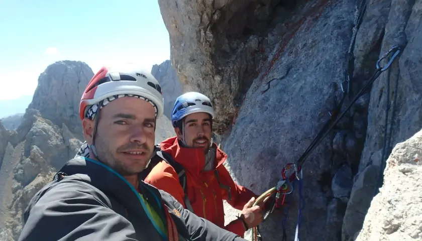 Dos escaladores sonríen en la cara del Naranjo de Bulnes, equipados con cascos y arneses, listos para la aventura.