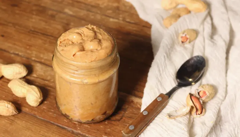 Barattolo di burro di arachidi fatto in casa, pronto per la tua prossima ricetta. Arachidi e cucchiaio su sfondo rustico.