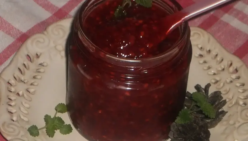 Słoik domowej frużeliny malinowej, z łyżeczką zanurzoną w gęstym, czerwonym musie. Udekorowana listkami mięty i szyszkami.