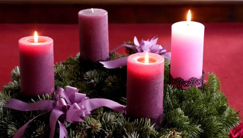 Drei von vier 3. Advent Kerzen brennen auf einem Tannenkranz mit lila Schleifen.
