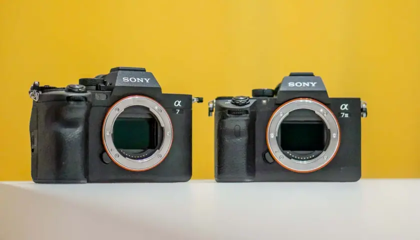 Dwa aparaty Sony Alpha, jeden z nich to sony a7 iiis, stoją obok siebie na białej powierzchni, na żółtym tle.