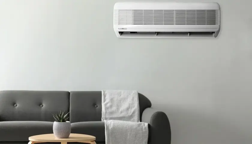 Unidad de aire acondicionado montada en la pared, cumpliendo la normativa aire acondicionado. Sofá gris con mantas y mesa auxiliar con planta.