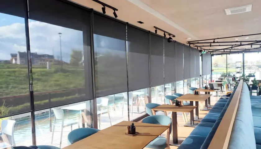 Nowoczesne wnętrze restauracji z widokiem na wodę, osłonięte eleganckimi roletami termicznymi na wymiar.