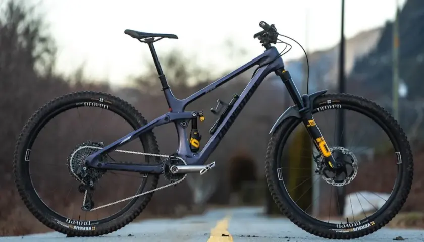A dark purple full-suspension mountain bike with yellow accents sits on a road. Its components suggest a focus on performance, hinting at a competitive mountain bike weight for agile trail riding.