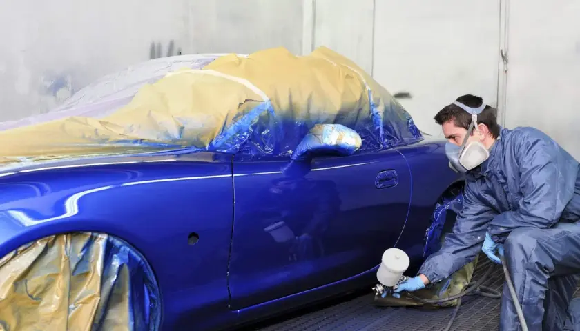 Un carrozziere con maschera spruzza vernice blu su un'auto coperta. Consulta il listino prezzi verniciatura auto per i costi.