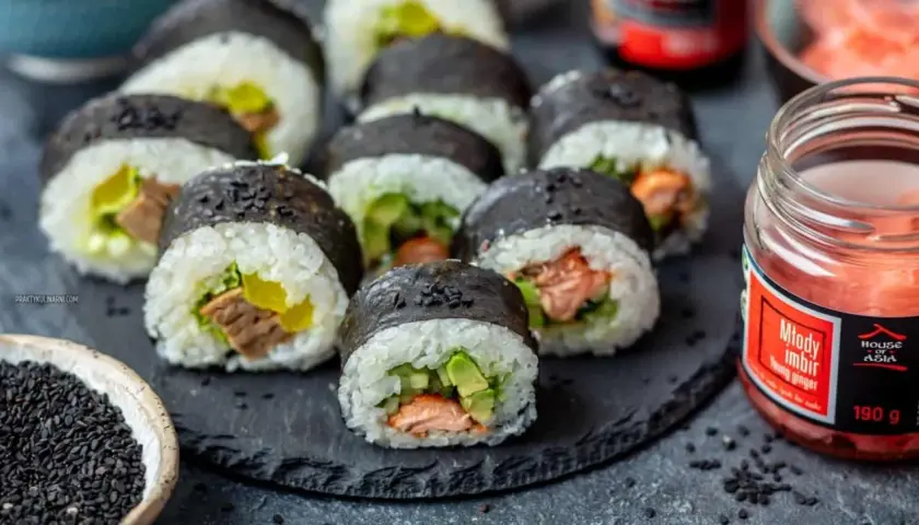 Gotowe do jedzenia rolki sushi z łososiem i awokado, obok słoiczek marynowanego imbiru i miseczka czarnego sezamu – idealne składniki do sushi.