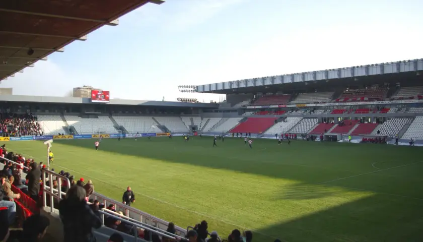 Widok na murawę i trybuny stadionu Cracovii. Na boisku trwa mecz, a trybuny zapełniają się kibicami.