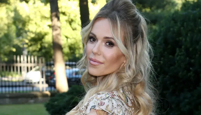 Doda IQ, blondynka w kwiecistej sukience, z burzą loków, uśmiecha się do obiektywu na tle zieleni.