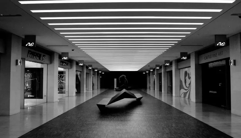 Un couloir de centre commercial, éclairci par des néons, avec une sculpture moderne au centre. Les boutiques sur les côtés sont sombres, créant un contraste saisissant.